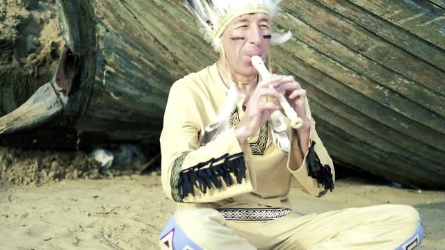 An Indian in a national costume plays a flute near a boat on the river bank 4k