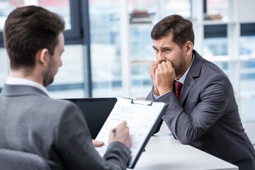 Nervous man in formal wear looking at businessman writing on clipboard during job interview, business concept