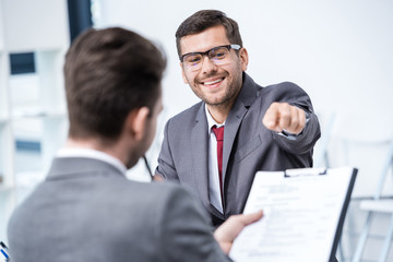 Smiling man in suit and eyeglasses pointing at clipboard at job interview, business concept