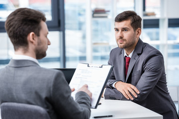 Serious man in formal wear looking at businessman with clipboard during job interview, business concept