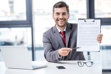 Fototapeta premium Handsome smiling businessman sitting at workplace and holding business contract, business concept