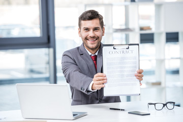 Handsome smiling businessman sitting at workplace and holding business contract, business concept