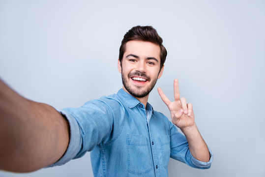 Handsome smiling young man is making selfie on smartphone and gesturing peace on light blue background