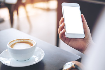 Mockup image of hands holding mobile phone with blank white screen and hot coffee cup on wooden table in cafe