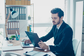 Concentrated handsome young man in black suit sitting at the table at office holding clipboard and...