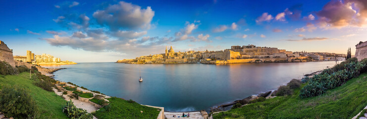 Fototapeta na wymiar Valletta, Malta - Panoramic skyline view of the ancient city of Valletta and Sliema at sunrise shot from Manoel island at spring time with sailing boat, blue sky, beautiful clouds and green grass
