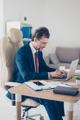 Side view of concentrated smiling man typing on laptop in office. He is in a suit, workplace in modern and nice