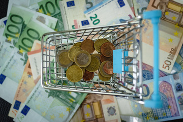 Euro coins in mini shopping cart and pile of euro banknote on background as shopping concept