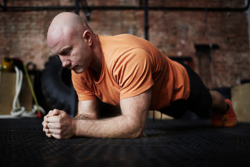 Bald middle-aged bodybuilder standing in plank during intensive workout in modern gym, full-length...