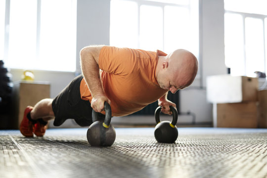 Bald bodybuilder looking down with concentration while doing plank exercise with kettlebells, full-length portrait