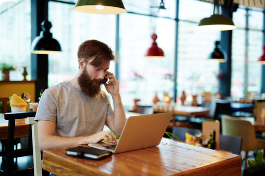 Busy bearded manager sitting in cyber cafe and talking to his business partner on mobile phone, modern laptop and notebook located on wooden table, waist-up portrait
