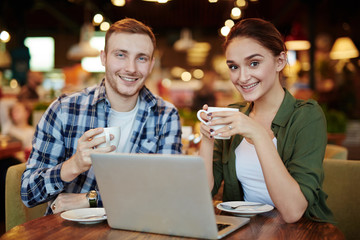 Two young friends posing for photography with toothy smiles while hanging out in small cozy coffeehouse, waist-up portrait