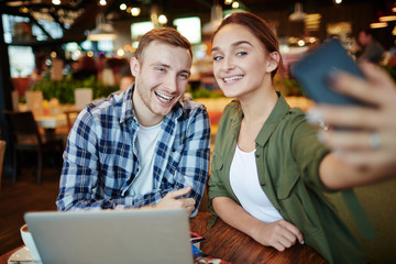 Lovely smiling couple taking selfie on smartphone while sitting in cozy small cafe, laptop, photos and cup of coffee located on wooden table