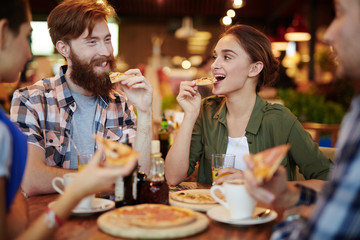 Portrait of pretty young woman biting into slice of pizza and looking at her smiling bearded friend while enjoying peaceful evening in cozy coffeehouse