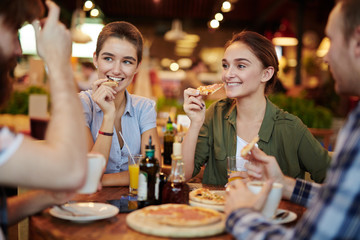 Red-haired bearded man telling interesting story to his friends while sitting in cozy pizza restaurant, two pretty women with wide smiles listening to him with attention, waist-up portrait