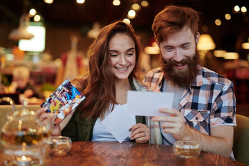 Loving young couple hanging out in cozy small cafe: they drinking fragrant tea and looking through printed photos, waist-up portrait