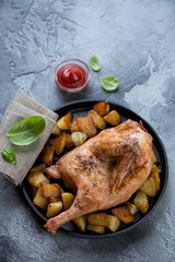 Oven baked half chicken and roasted potato wedges in a frying pan, vertical shot on a grey stone surface