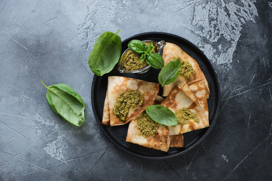 Stuffed crepes topped with basil pesto sauce. Top view on a dark grey stone surface, horizontal shot
