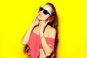 Beautiful young sexy girl dj in blue sunglasses and red striped T-shirt laughing and having fun and listening to music in red headphones on a yellow background