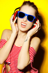 Beautiful young sexy girl dj in blue sunglasses and red striped T-shirt laughing and having fun and listening to music in red headphones on a yellow background