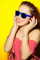 Beautiful young sexy girl dj in blue sunglasses and red striped T-shirt laughing and having fun and listening to music in red headphones on a yellow background