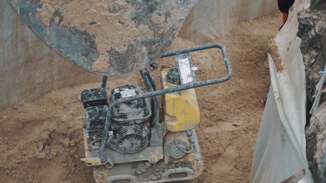 Slowmotion close up excavator laddle pulling plate compactor down on sand in ditch, worker unstraped it