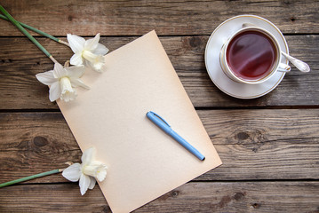 A piece of paper with pen and white daffodils and a cup of tea on wooden background