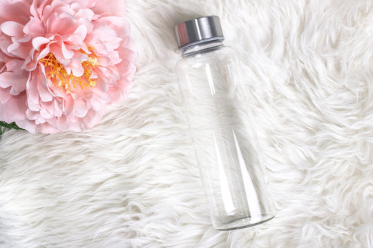 Styled Mockup Of Clear Plain Glass Water Bottle On White Faux Fur With A Flower. Custom Gifts.