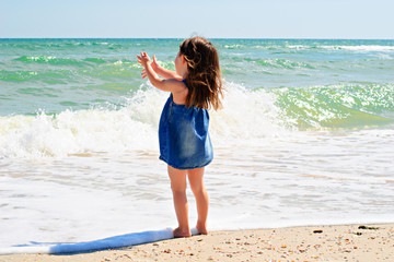 Cute little girl in denim dress standing alone on the beach saying hello to the sea