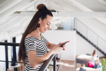 Young beautiful woman with brown hair is standing indoors and holding a smartphone. A charming brunette female in a cafe surfing the net with a mobile phone.