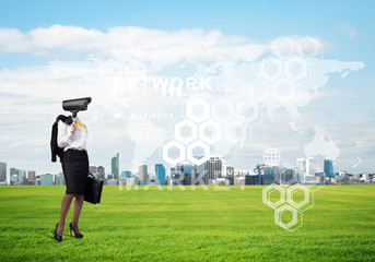 Camera headed woman standing on green grass against modern cityscape