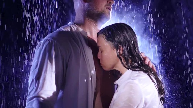 The woman kisses the man's chest with an unbuttoned white shirt under a big rain