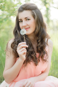 Portrait of a beautiful young girl in a tender dress. Sits among the flowering spring trees in the park in the open air. In the hand holds a dandelion. Fine art