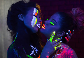 Sexy lesbian fashion models kissing in uv neon light with fluorescent glowing Body Art make-up ....