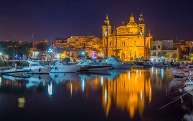 Fototapeta na wymiar Msida, Malta - The beautiful Msida Parish Church with yachts and boats and reflection on the water by night