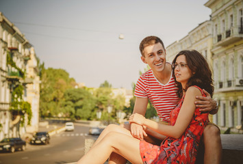 Cute young smiling couple in love hugging, sitting outdoors at green city street, summertime. Girl is wearing glasses of her boyfriend