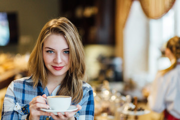 Young blonde woman enjoying coffee or chocolate in cozy cafe. Female holding cup of cappuccino. Blurred interior behind the model.
