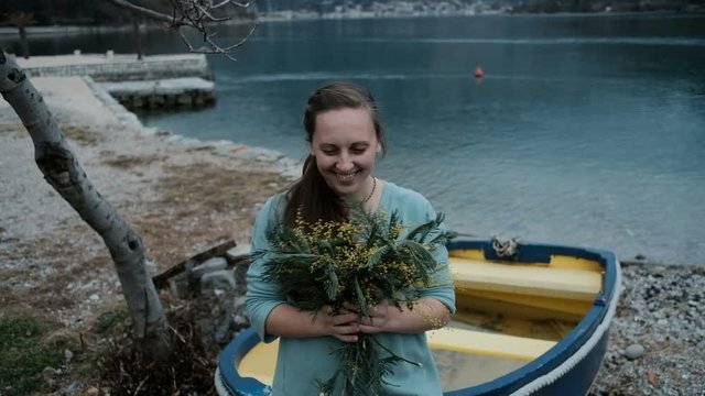 Adult Caucasian woman is happy receive bouquet of mimosa near lake. young fair-haired lady with hairstyle based on tress fixed by white scrunchy in blue dress or blouse, small necklace, gold ring hold