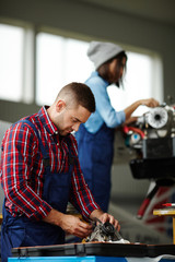 Two modern mechanics, man and woman, repairing engine parts in workshop, both wearing workers uniform and overalls