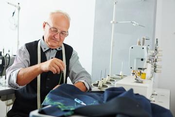 Fototapeta premium Portrait of pleasing old man working in tailoring studio making clothes at sewing machine and hand stitching cloth