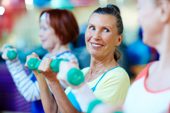 Smiley mature woman with dumbbells and her friends exercising in gym