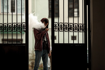 Vape man. Photo of a handsome young white guy in the hood vaping and letting off steam from an electronic cigarette near a vintage metal gate. Lifestyle.