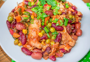 Closeup homemade stewed beans with chicken meat green peas and vegetables served with cut green onion and parsley on a white plate on wooden table