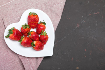 fresh red strawberries on white heart shaped plate on napkin on black tabletop, berries top view concept