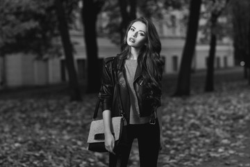 Beautiful stylish girl in gray pullover, black jeans and leather jacket standing and posing in autumn park. Pretty young woman with long curly brunette hair and red lips holding handbag