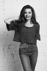 Young beautiful gorgeous female model in blue jeans and purple pullover posing against white brick wall. Stunning glamorous happy joyfull smiling girl with long curly hair