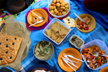 Unrecognizable people eating various picnic food: roasted vegetables salad, baba ghanoush, gluten-free crackers, foccacia bread, gluten-free and sugarfree dates cake. Top view. 
