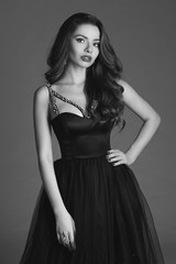 Fototapeta premium Young beautiful woman standing and posing in black ball gown on white gray background. Fashion style portrait of girl with long curly brunette hair