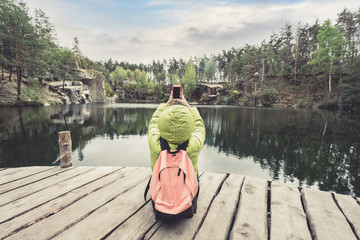 A female traveler in green jacket and pink-brown backpack is sitting on a wooden pier near a beautiful lake surrounded by stone rocks in the middle of a pine forest and taking photos on mobile phone.