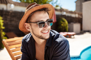 Man in sunglasses and hat smiling, sitting near swimming pool.